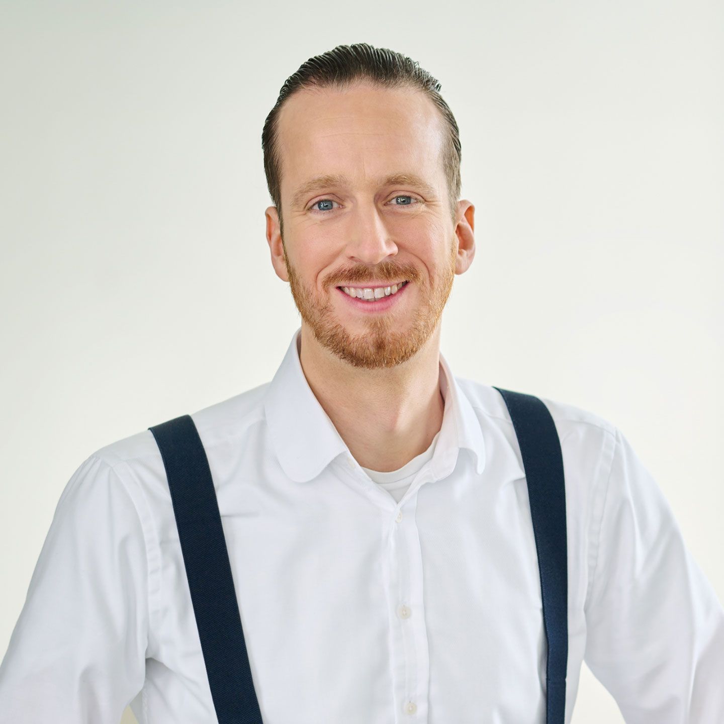 Sebastian Pollmer, Mann steht in einem weißen Hemd und blauen Hosenträgern in einem hellen Raum. 