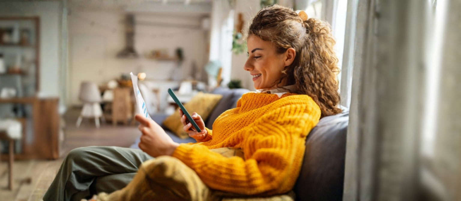 En smilende ung kvinde med krøllet hår, iført en hyggelig gul sweater, sidder i en sofa i sin stue og kigger på sin smartphone, mens hun holder et papirdokument.