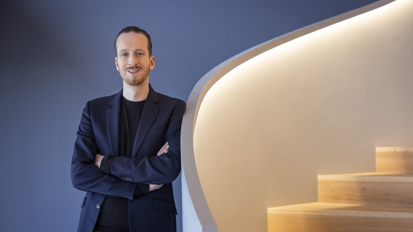 Sebastian Pollmer a male in a blue suit leans against a wall near to a white warm lightended stair