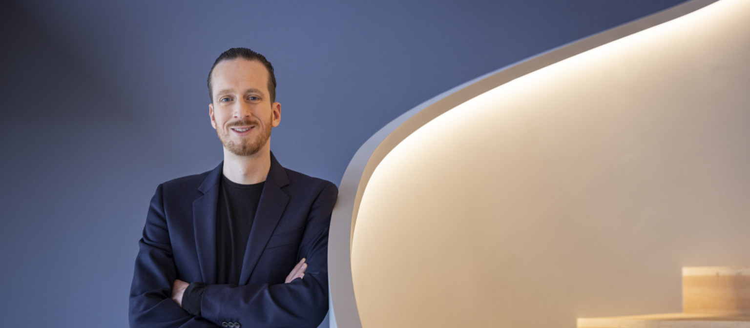 Sebastian Pollmer a male in a blue suit leans against a wall near to a white warm lightended stair