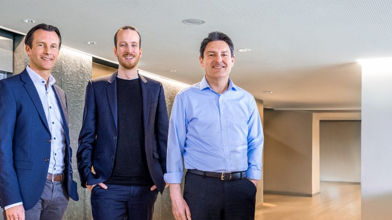 Three male members of the EOS Group’s Board of directors are standing in business attire in front of elevators in a modern setting.