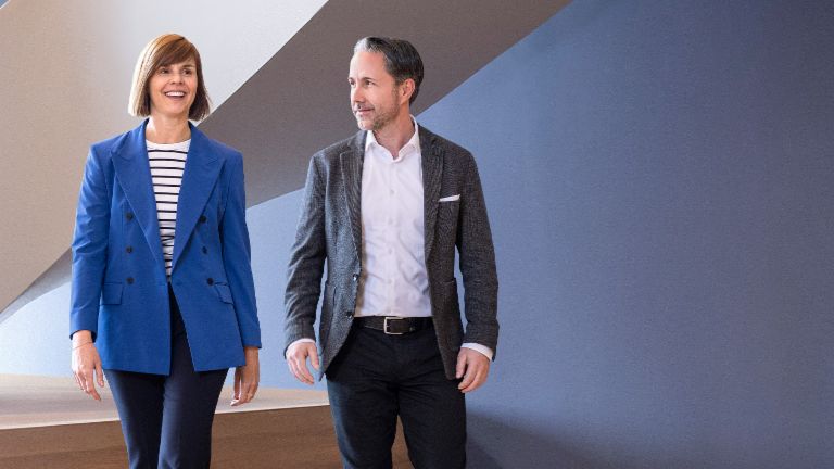 The CEO in a gray blazer and the CFO of the EOS Group in a blue blazer walk through a modern office. The space features large windows, a curved wall, and stylish hanging lamps.