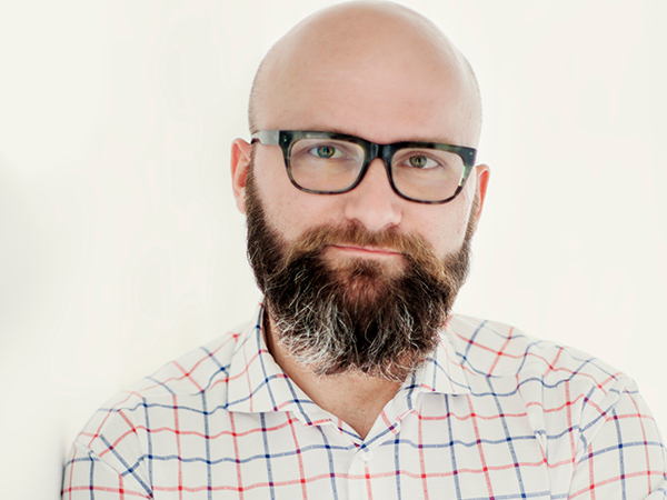 Dr. hab. Tomasz Piekot, wearing a checked shirt, leans against a wall with his arms crossed