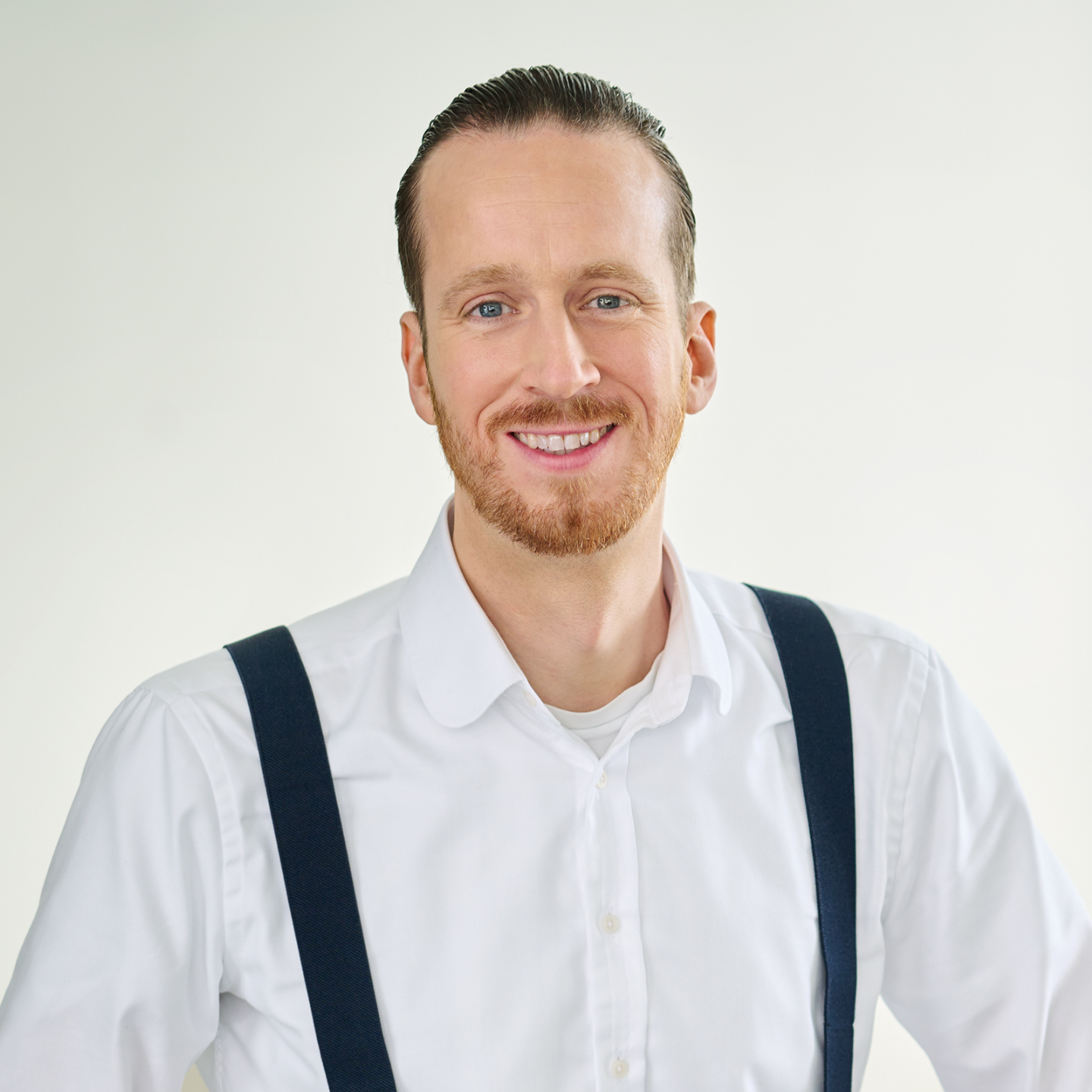 Sebastian Pollmer, man standing in a white shirt and blue braces in a bright room. 