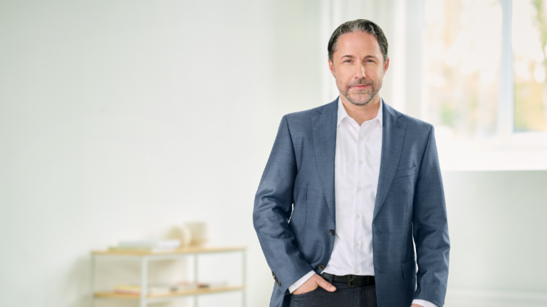 Marwin Ramcke, CEO, standing in a blue suit, with his right hand in his trouser pocket, in a bright room. 